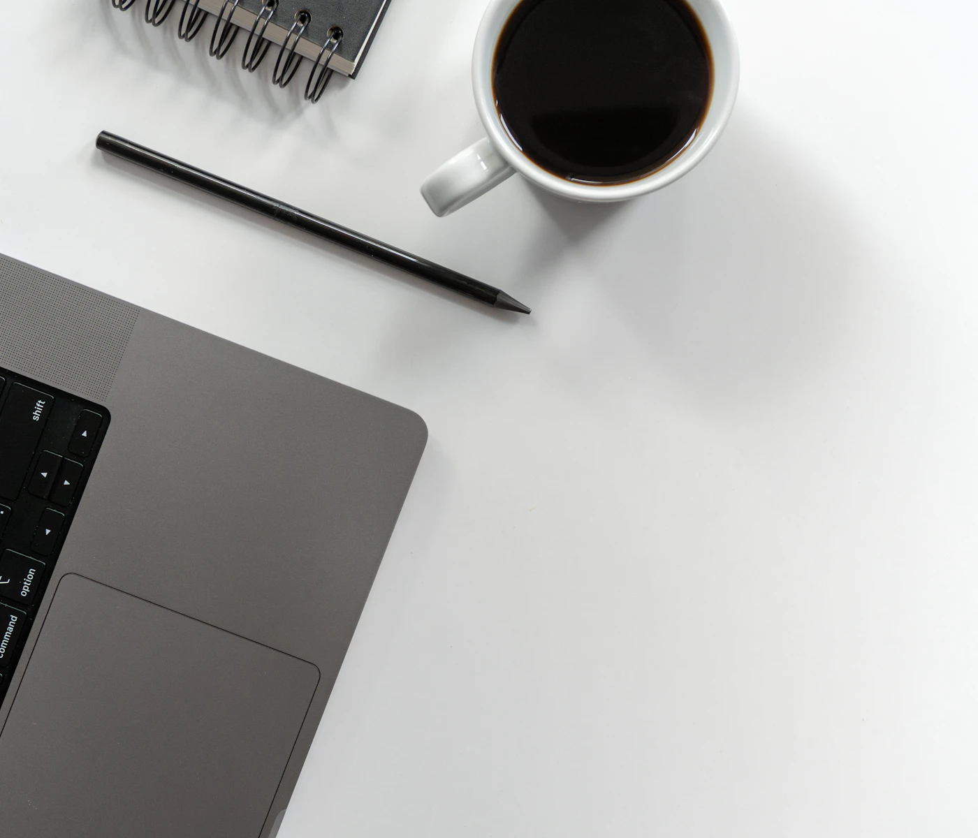 Top-down view of a laptop corner, a black pencil, a spiral notebook and a cup of black coffee on a white desk